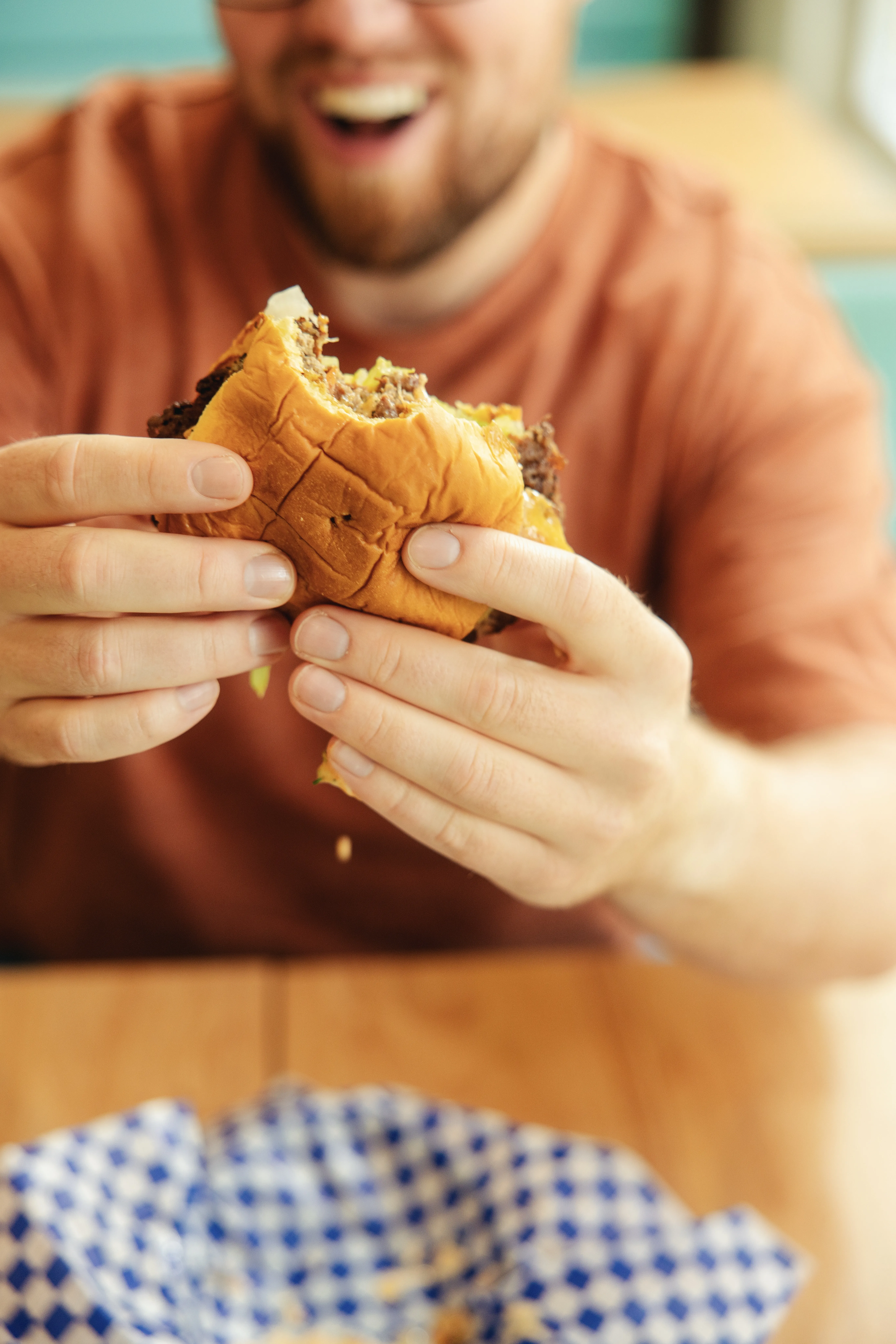 A man holding a burger with a bite taken out of it, the focus is on the food as a drop of sauce falls from the fresh smashburger.